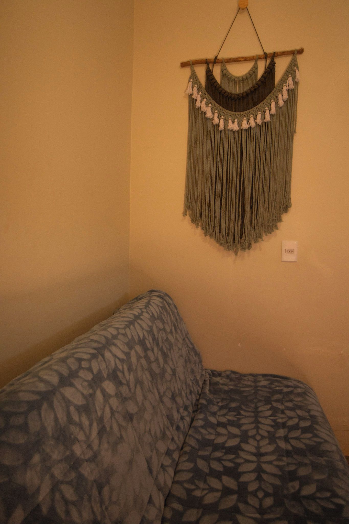 Een kamer met een bed met een bladmotiefdeken, een macramé kunstwerk aan de muur en een stopcontact.