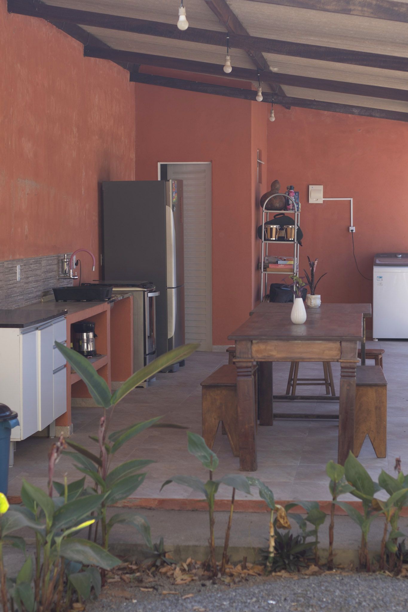 A compact kitchen with a refrigerator, stove, and sink. There is a dining table with benches and a potted plant.