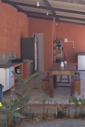 Cocina compacta con refrigerador, estufa y lavamanos. Hay una mesa con bancos y una planta en maceta.