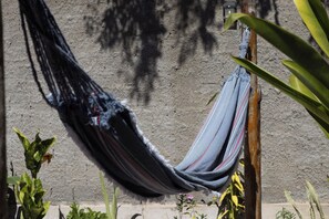 Una hamaca colgada entre dos postes de madera, con un muro de hormigón al fondo y algo de vegetación alrededor.