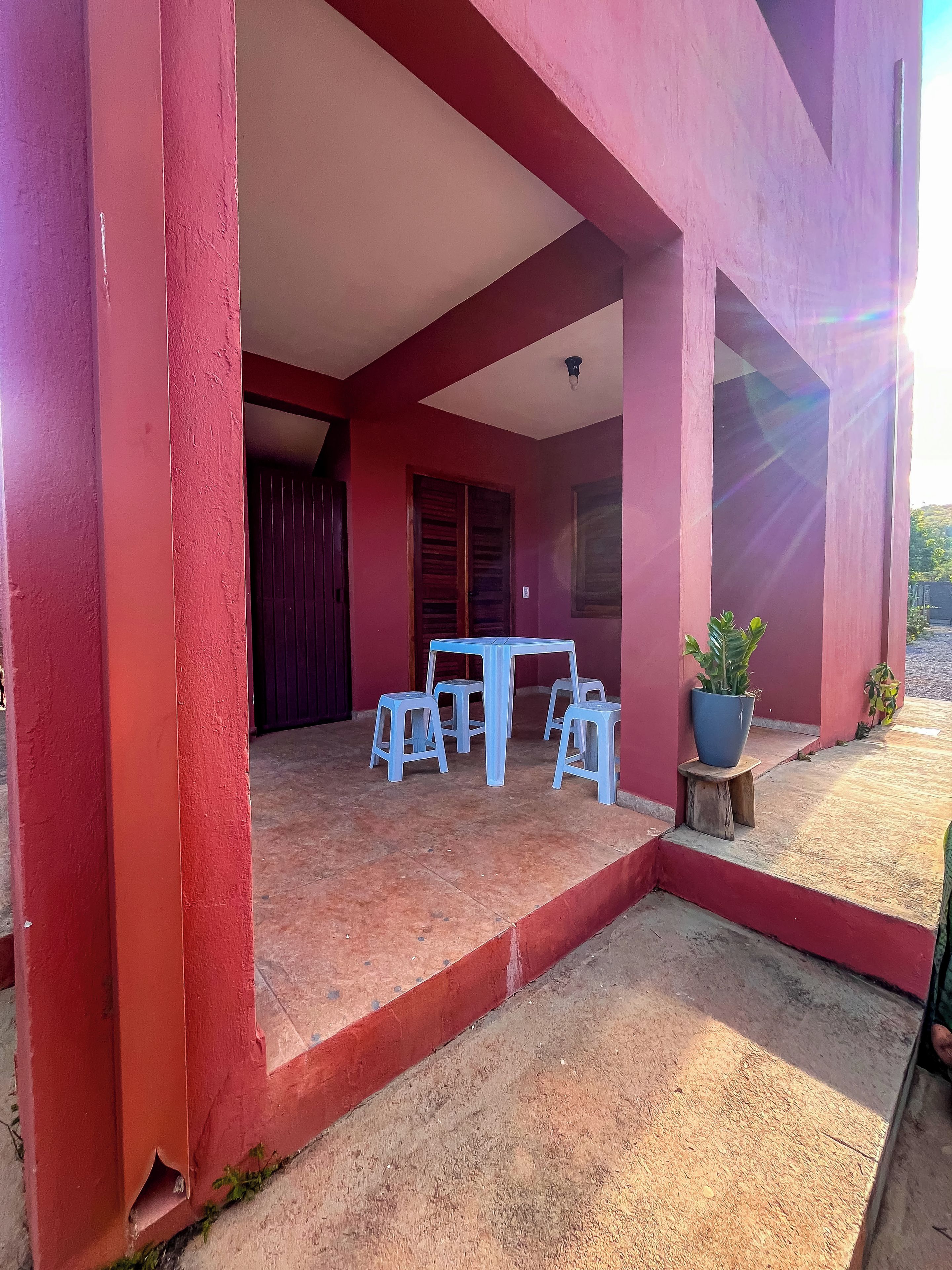 Un edificio rojo con un patio techado que cuenta con una mesa y cuatro taburetes, una planta en maceta y un taburete de madera.