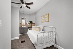 Ein Schlafzimmer mit einem weißen Bett, einem Deckenventilator, einem Badezimmer mit Waschbecken und Spiegel sowie zwei gerahmten Bildern an der Wand.