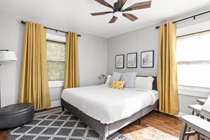 Un dormitorio con cama, ventilador de techo, ventana con cortinas, mesita de noche y lámpara.