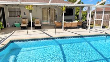 A swimming pool with a covered patio area, outdoor furniture, and potted plants.