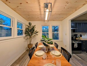 A dining room with a wooden table set for six, flanked by windows and a potted plant. - Boutique Farm Stay | Highland Cow * Horses * Bunnies (Cresco)