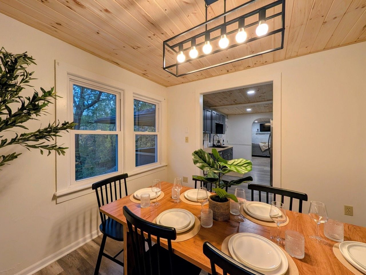 Un comedor con una mesa de madera dispuesta para cuatro personas, rodeada de sillas, con vistas a la cocina y un ventanal grande.