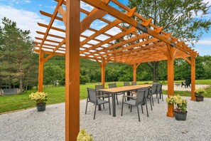 A wooden pergola structure with a table and chairs underneath, surrounded by gravel and potted plants. - Boutique Farm Stay | Highland Cow * Horses * Bunnies (Cresco)