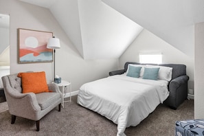 A bedroom with a bed, a chair, a lamp, and a painting on the wall. - The Cockatoo - Two Queen Beds (Indianapolis)