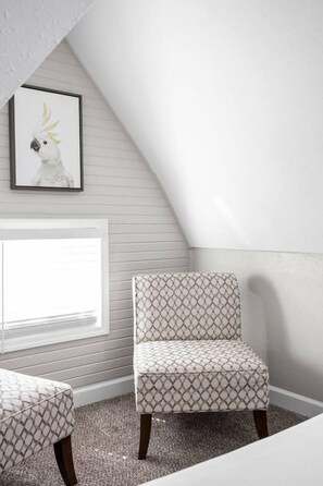 A room with a sloped ceiling, a window with blinds, a patterned chair, and a framed picture on the wall. - The Cockatoo - Two Queen Beds (Indianapolis)