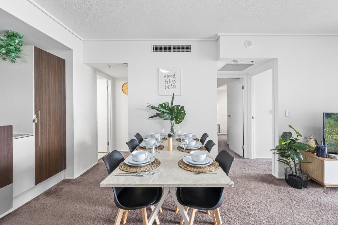 Un coin repas avec une table dressée pour quatre personnes, entourée de chaises noires, dans une pièce aux murs blancs et à la porte en bois.
