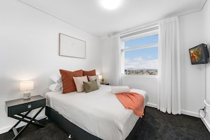 Une chambre d’hôtel avec un lit, une table de chevet, une lampe, une fenêtre donnant sur un beau paysage et un tableau au mur.