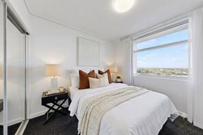 Ein Hotelzimmer mit Bett, Nachttischen, Lampen, einem Fenster mit Blick auf die Stadt und einem gerahmten Kunstwerk an der Wand.