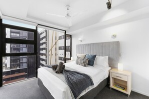Un dormitori modern amb un llit gran, una tauleta de nit, un ventilador de sostre i vista a un edifici a través d'una finestra gran.