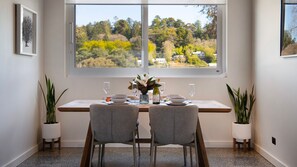 A dining area with a table set for two, two chairs, a vase with flowers, and a window with a view of trees and houses. - Marlowe sleek sustainable townhouse (Daylesford)