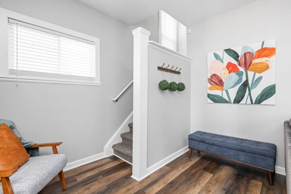 A room with a grey wall, wooden flooring, a grey sofa, a blue bench, a staircase, and a wall-mounted artwork. - College Ave 6 Bedroom House Sleeps 14 (Indianapolis)