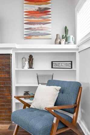 A room with a blue armchair, a white cushion, a fireplace, a shelf with decorative items, and a wall-mounted abstract painting. - College Ave 6 Bedroom House Sleeps 14 (Indianapolis)
