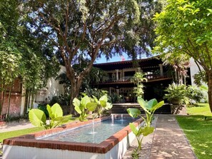 Une cour avec une piscine rectangulaire, entourée de grands arbres et d’un bâtiment doté d’un balcon.