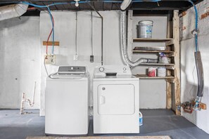 A laundry room with two washing machines, a water heater, and a shelf with cleaning supplies. - Earthy Escape - 5 min to downtown (Indianapolis)