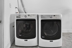 Deux machines à laver à chargement frontal se trouvent dans une buanderie, avec un tuyau visible et une porte sur le côté gauche.