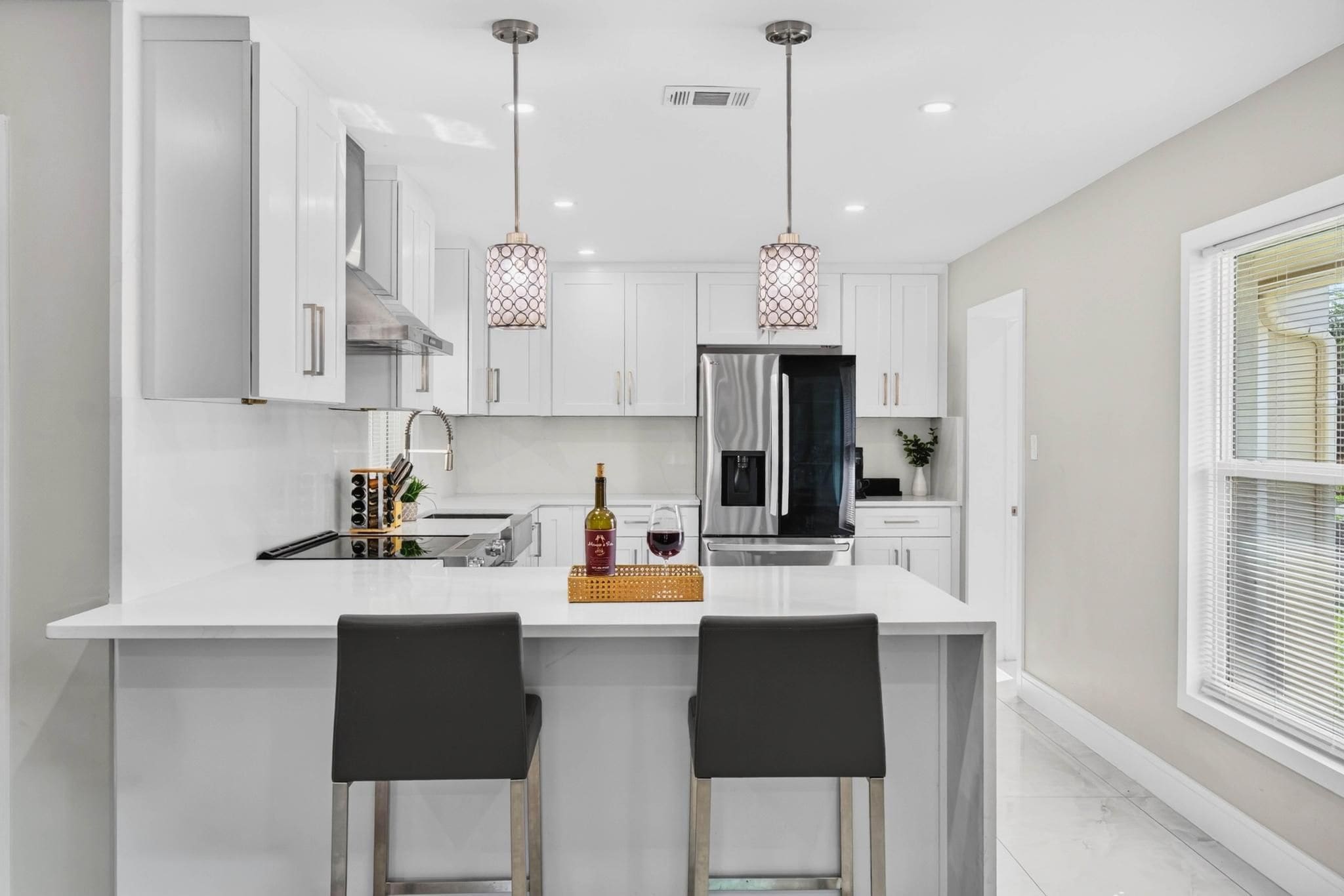 A modern kitchen with a central island, two pendant lights, and a large window with blinds.