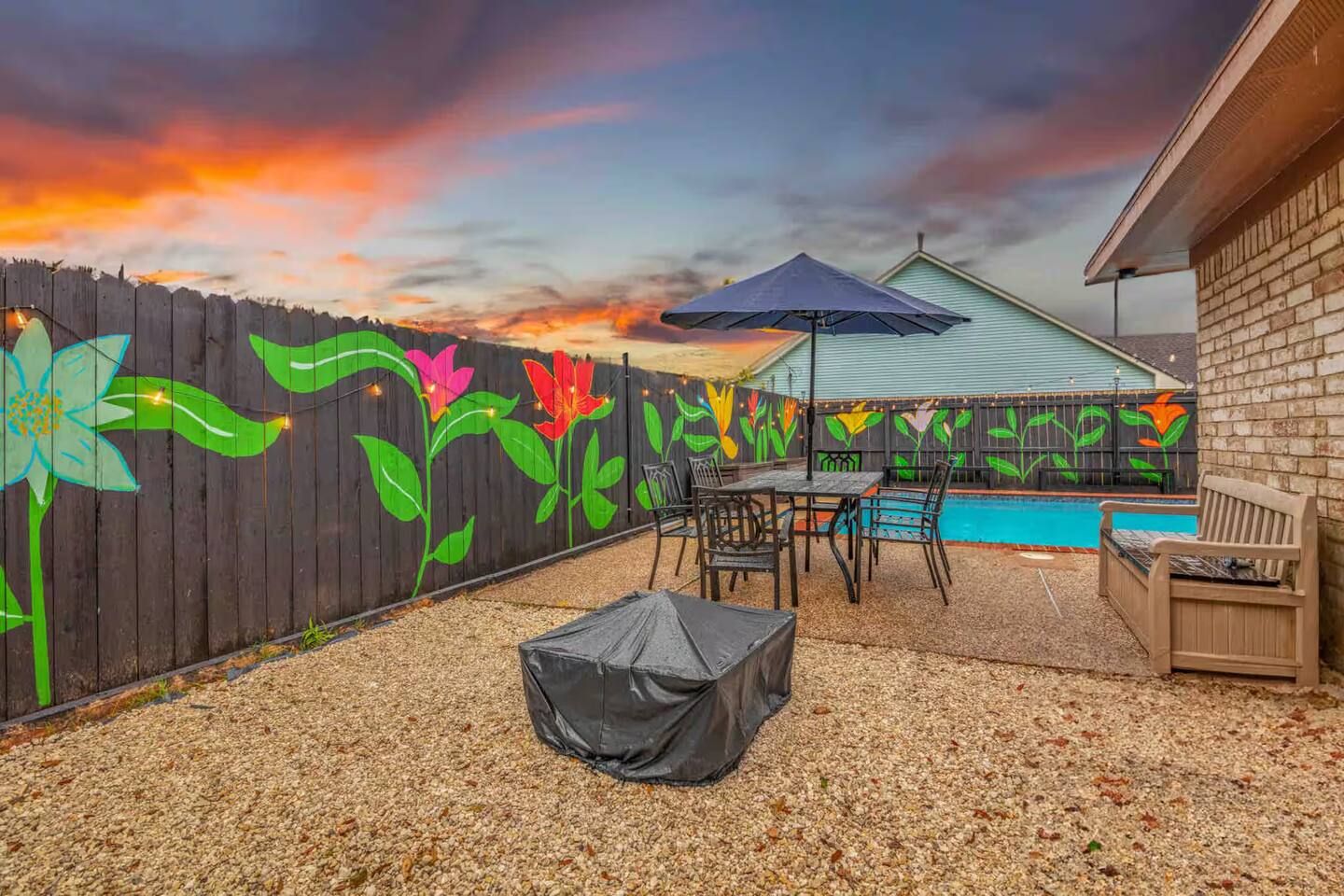 Un jardin avec une piscine, une table de patio couverte et une clôture ornée de peintures murales représentant des fleurs aux couleurs vives.