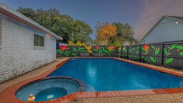 Une piscine avec un jacuzzi, entourée d’une terrasse en briques et d’une clôture en bois ornée de peintures murales florales aux couleurs vives.