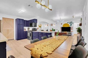 A modern kitchen with dark blue cabinets, a large island, and a long dining table with a woven placemat. - Boho Retreat 5BR Sleeps14 Pool 2TVs DeerPark (Deer Park)