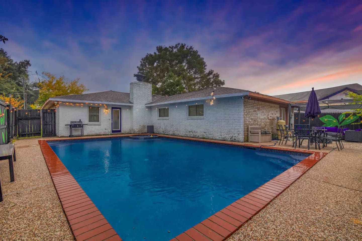 A swimming pool with a brick border, a small house with a chimney, and outdoor furniture.