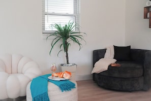 A living room with a white armchair, a dark sofa, a small table with a glass of drink and fruit, and a potted plant. - TPA Fun Compound by Airport / Restaurants (Tampa)