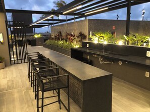 An outdoor bar area with seating and a counter, surrounded by plants and under a covered structure. - New Listing (João Pessoa)