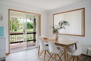 A dining area with a wooden table, white chairs, a vase with flowers, and a window with blinds. - Paradise Lake Conjola Park - Wake up to ocean views (Conjola Park)