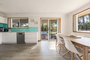 A modern kitchen with a dishwasher, a dining area with a table and chairs, and a sliding glass door leading to an outdoor deck. - Paradise Lake Conjola Park - Wake up to ocean views (Conjola Park)