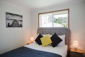A bedroom with a bed, pillows, a nightstand, a lamp, a window with a view, and a painting on the wall. - Paradise Lake Conjola Park - Wake up to ocean views (Conjola Park)