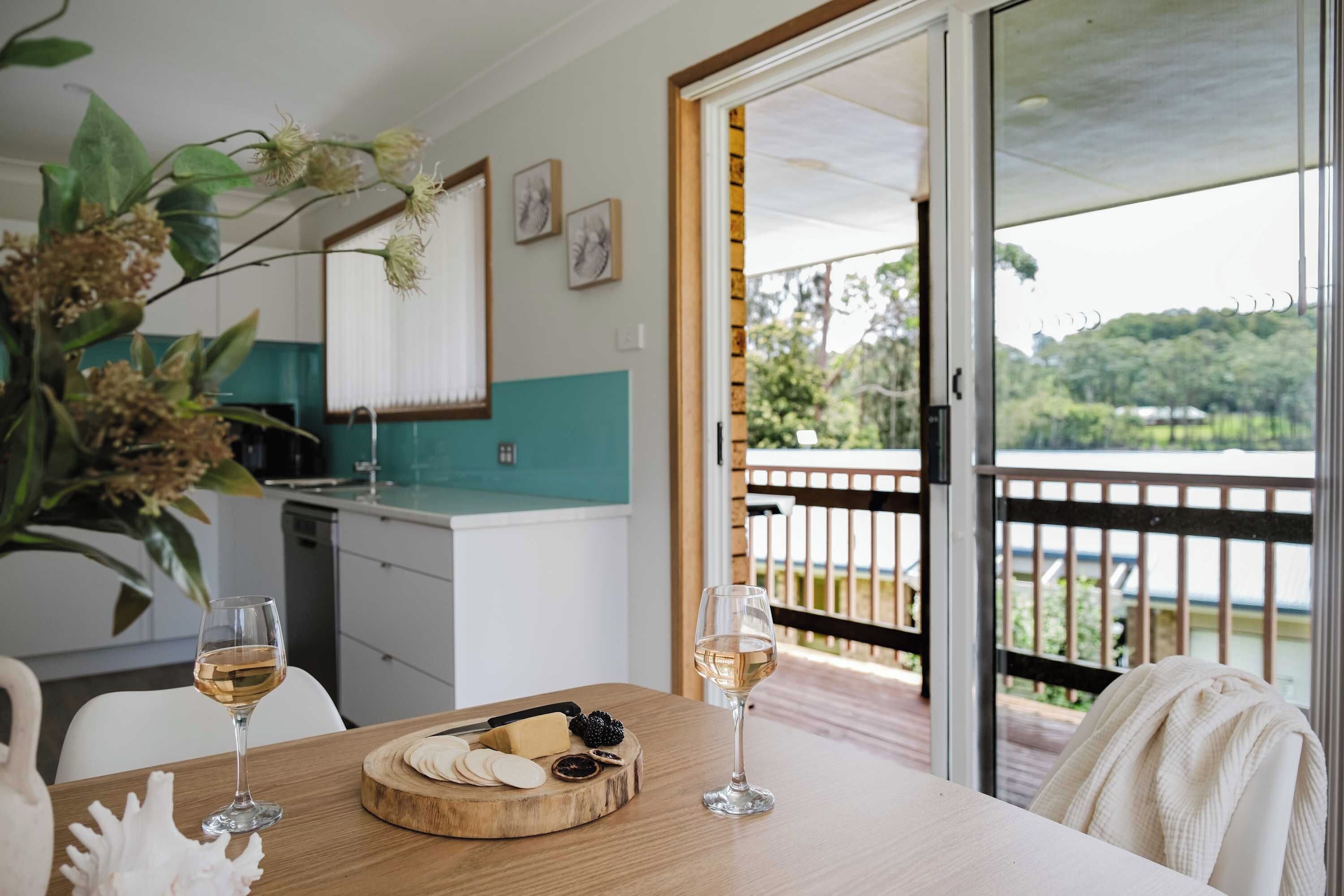 A dining area with a wooden table, two wine glasses, a cheese board, and a view of a balcony with a railing.