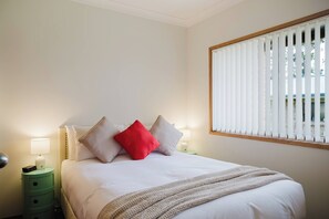 Un dormitorio con cama, almohadas, lámpara de noche, mesita de noche y ventana con persianas.