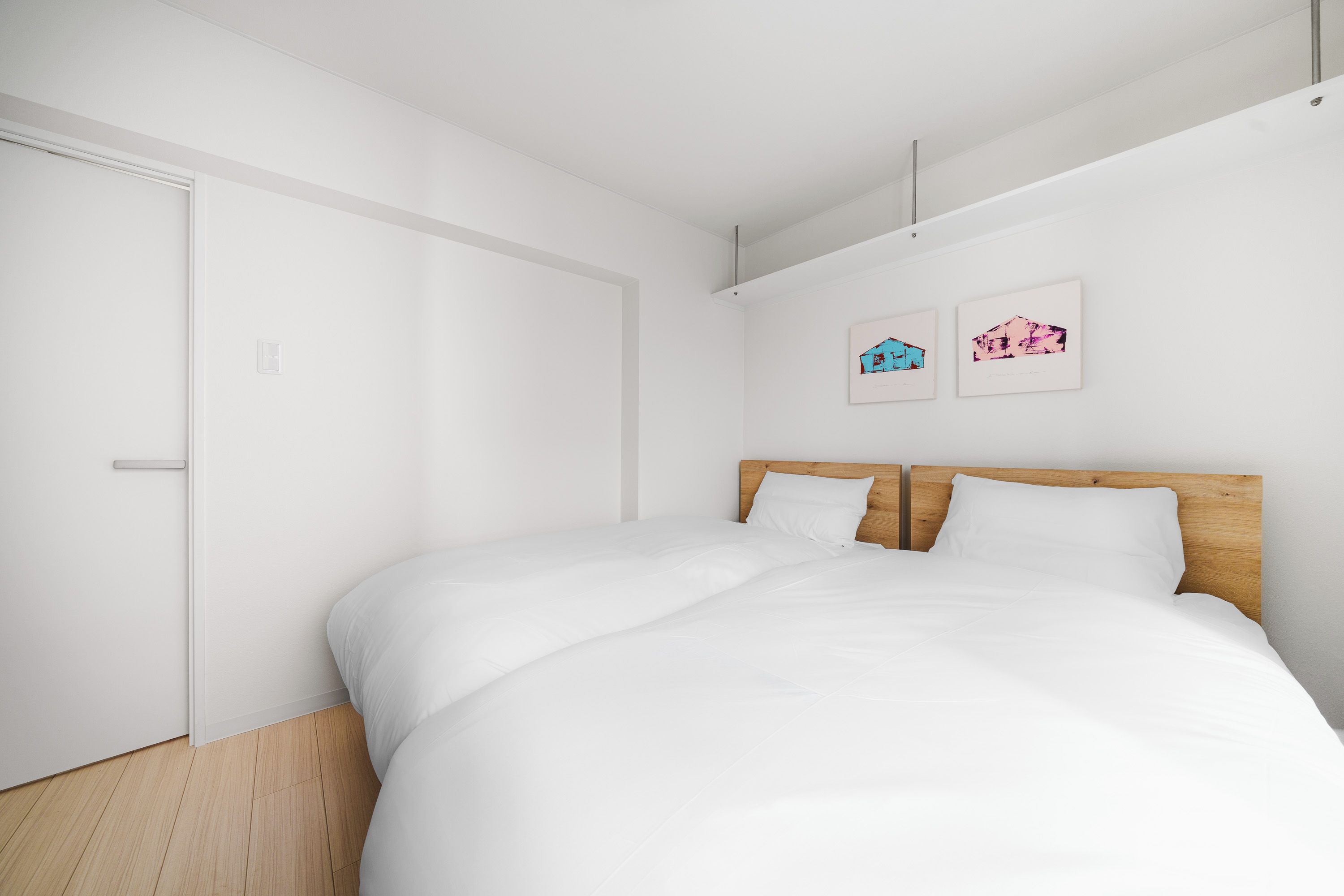 A minimalist bedroom with a wooden headboard, white bedding, and two wall-mounted artworks.