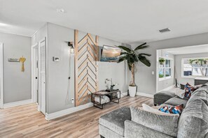 A living room with a grey sofa, a wooden herringbone wall, a large potted plant, and a flat-screen TV mounted on the wall. - 🌴Palm Breeze Retreat- Home with a pool, hot tub & game room near Clearwater Beach! (Largo)