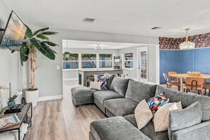 A modern living room with a grey sectional sofa, a dining area with a wooden table and chairs, and a kitchen visible through an open door. - 🌴Palm Breeze Retreat- Home with a pool, hot tub & game room near Clearwater Beach! (Largo)