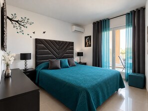 Une chambre moderne avec un grand lit, deux tables de chevet, un vase avec des fleurs, un climatiseur mural et une vue sur l’extérieur grâce à une porte coulissante.