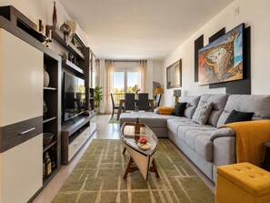 Un salon moderne avec un canapé gris, une table basse et un téléviseur. On y trouve également un coin repas avec une table et des chaises, ainsi qu’un balcon avec vue.