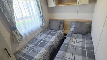 A compact room with two single beds, a small wooden shelf, and a window with curtains.