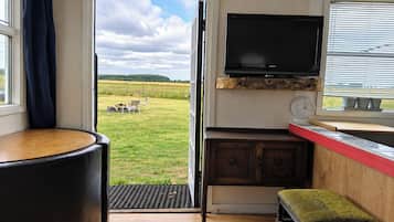 A room with a television mounted on a wall, a wooden table, a bench, and a view of a grassy field through a window.