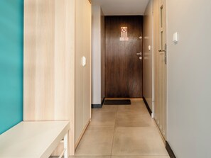 A hallway with a wooden door at the end, a light-colored bench on the left, and a wall-mounted light fixture on the right. - Legionów Central Studio #4 (Łódź)