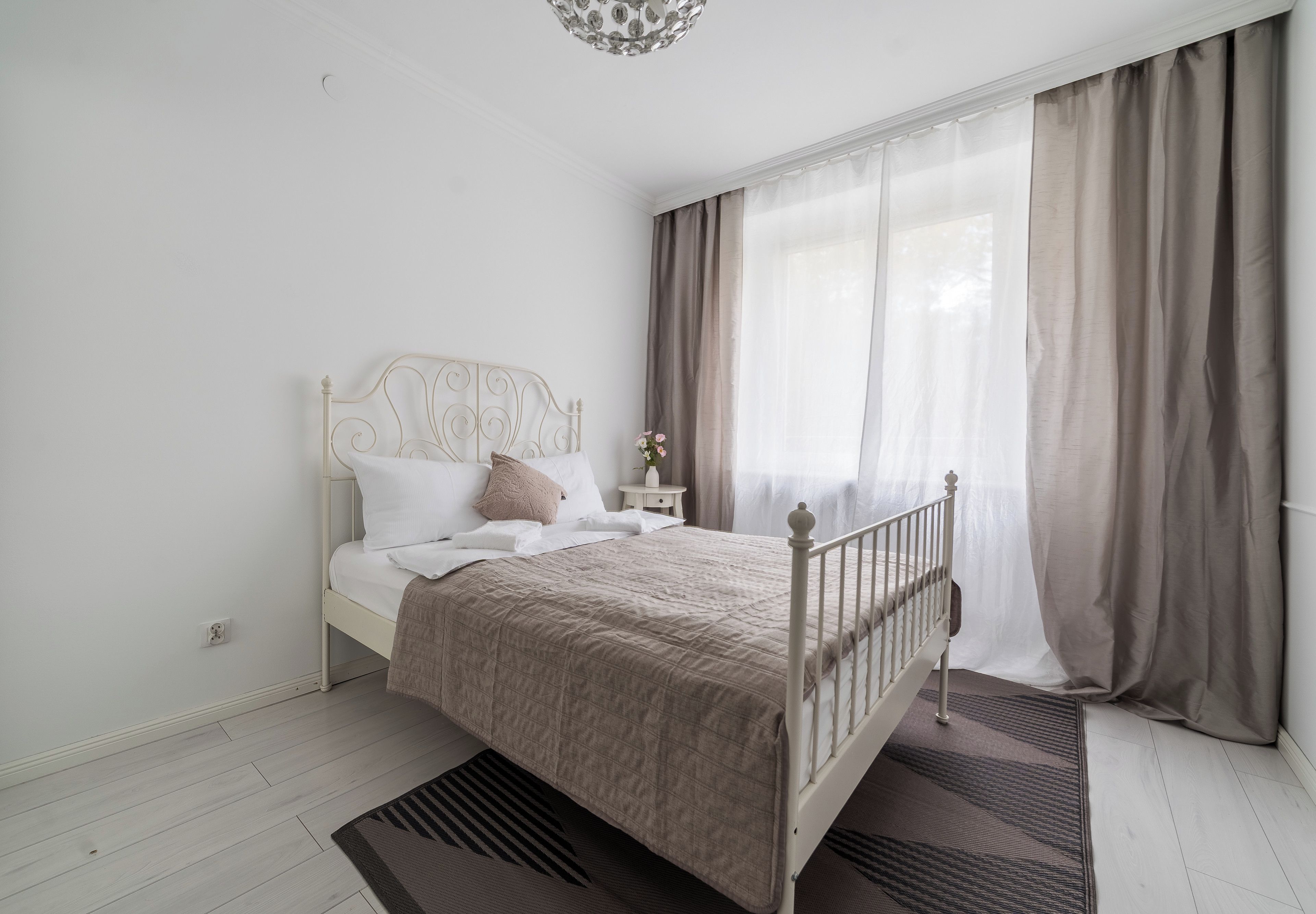 Ein Schlafzimmer mit einem Bett, einem Nachttisch, einer Vase mit Blumen und einem Fenster mit Vorhängen.