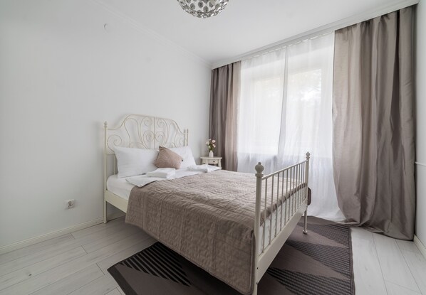 Ein Schlafzimmer mit einem Bett, einem Nachttisch, einer Vase mit Blumen und einem Fenster mit Vorhängen.