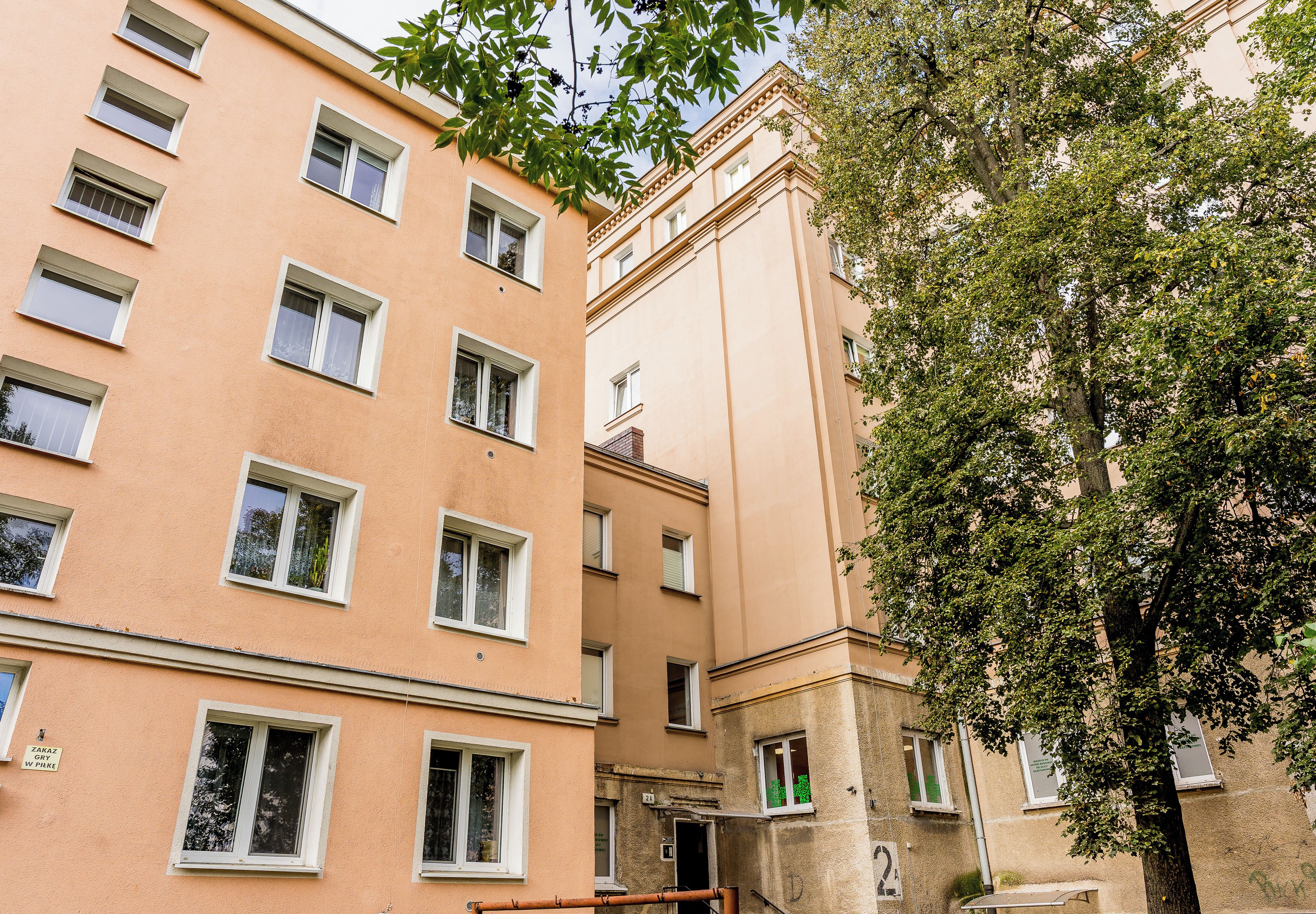 Ein mehrstöckiges Gebäude mit einer beigefarbenen Fassade und mehreren Fenstern, im Vordergrund steht ein Baum.