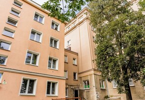 Ein mehrstöckiges Gebäude mit einer beigefarbenen Fassade und mehreren Fenstern, im Vordergrund steht ein Baum.