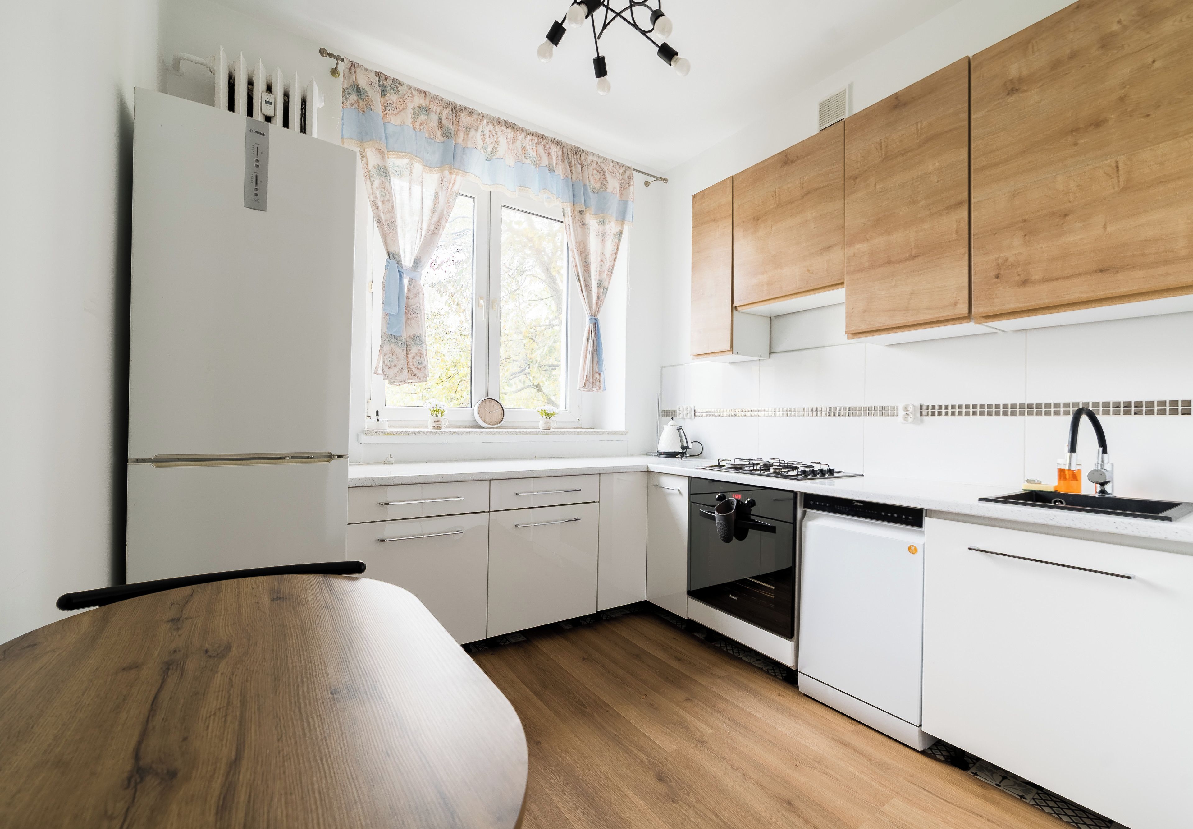 A modern kitchen with white cabinets, a wooden dining table, and a window with curtains.