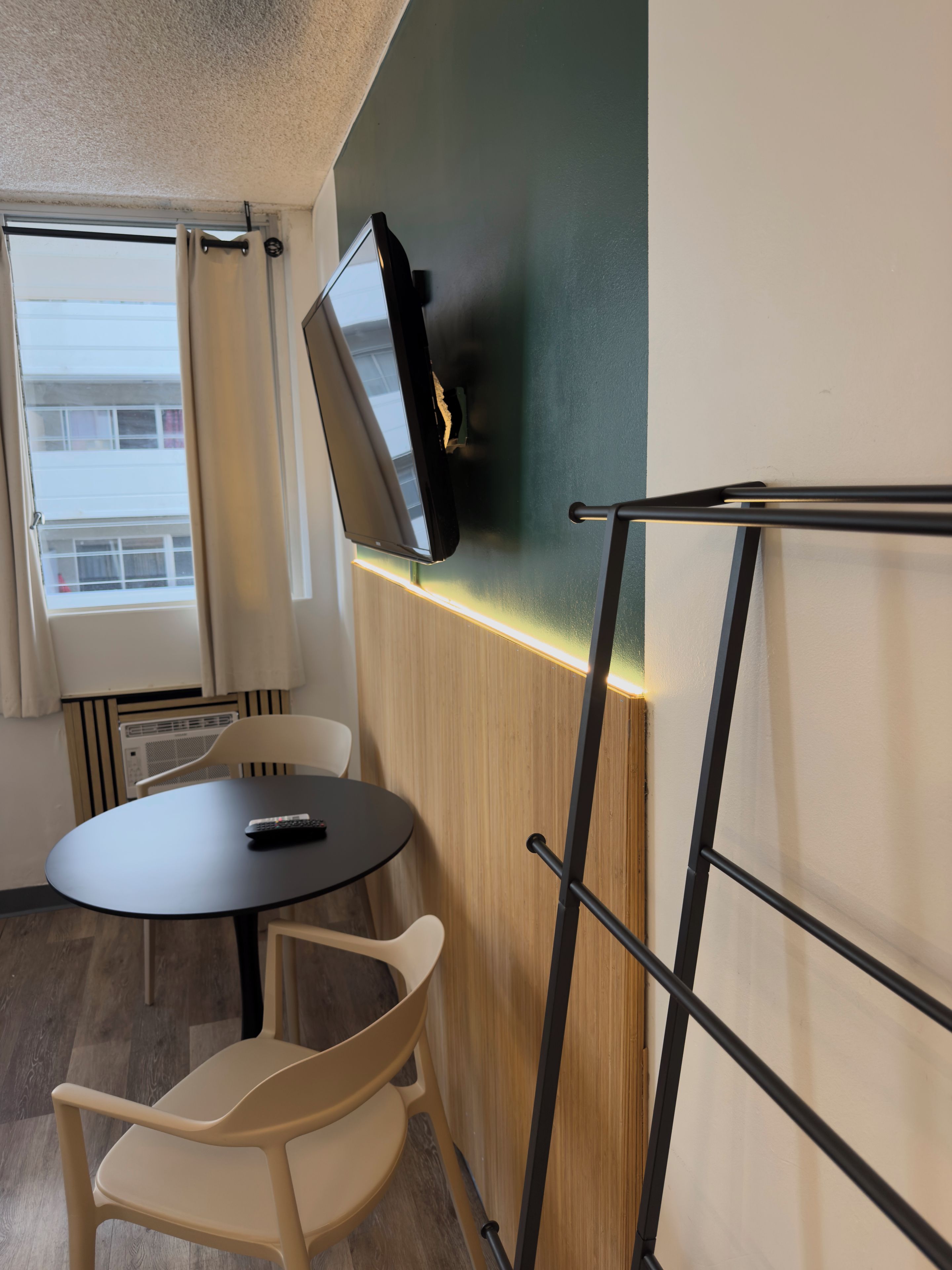 A compact room with a television mounted on the wall, a small round table, a chair, and a window with curtains.