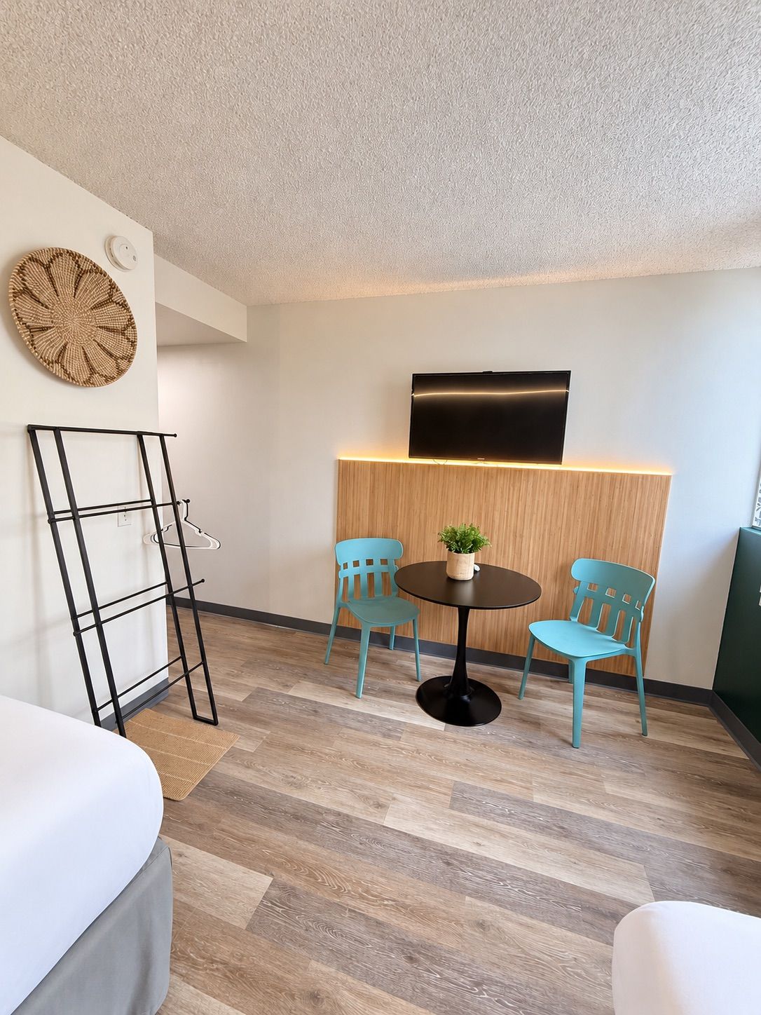 A modern room with a bed, a small round table with two chairs, a TV mounted on a wooden panel, and a wall-mounted shelf.